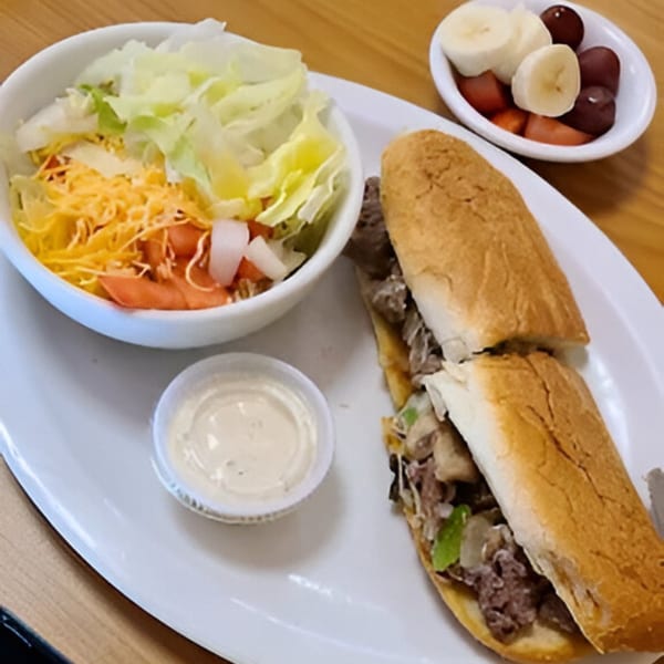 Cheesesteak sandwich with a side salad, dressing, and a small bowl of fruit.